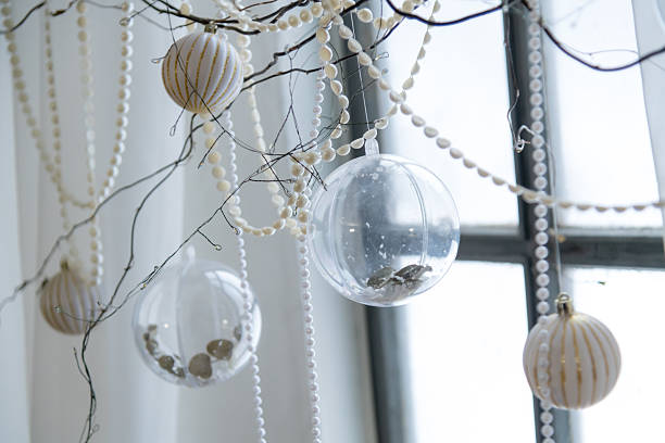 Decorations hang from branches in a bright room with natural light. Clear and white ornaments suspend in the air. Beads and strings create a festive atmosphere around the window.