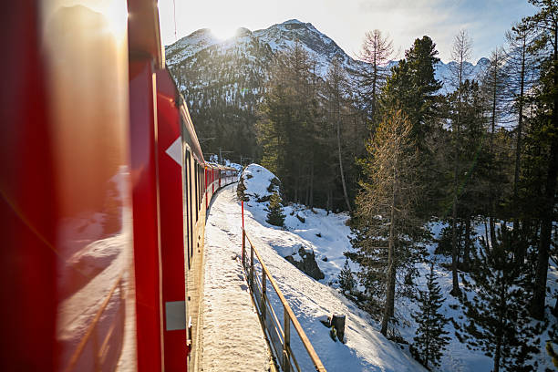 Red train moving through snowy winter mountains and pine forest with sunlight overhead