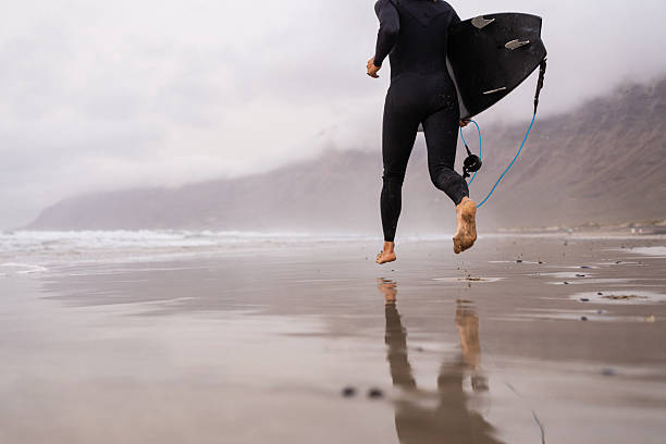Handsome young male surfer in a wetsuit comes out of the water to the shore past wooden posts and holds a surfboard in his hands