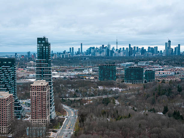 A scenic urban landscape capturing modern glass residential skyscrapers in North York, Toronto. The composition features a railway line cutting through a wintry valley in the foreground, leading the eye toward the distant Toronto downtown skyline and the iconic CN Tower under an overcast sky. This image illustrates urban development, modern architecture, and the expansive city layout of the Greater Toronto Area (GTA) during the winter season.