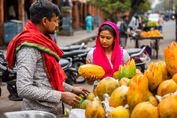 A street fruit stall scene where a young woman in a bright pink headscarf inspects a large papaya held in her hands while a male vendor in a patterned kurta and red shawl reaches for more fruit. Several ripe papayas — some with decorative carved tops — fill the foreground, with parked scooters and other market stalls blurred in the background. The image captures a candid moment of everyday market shopping and colorful produce on a busy street.
