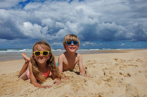 Two children wearing colorful sunglasses are happily playing in the sand on a beach, with gentle waves and a dramatic cloudy sky creating a beautiful summer scene by the ocean