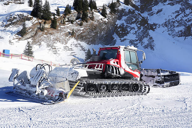 Red snow groomer vehicle stands parked on snowy mountain ridge under dramatic cloudy sky. Snowcat captured against scenic alpine backdrop with snow-covered peaks