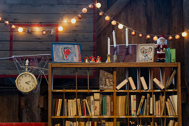 Rustic bookshelf with warm string lights, books and framed art, candles and Santa figure, hanging wall clock in cozy wooden workshop or barn interior
