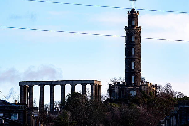A tall, dark stone tower stands on a hill beside a colonnaded structure under a pale blue sky  in Edinburgh, Scotland.