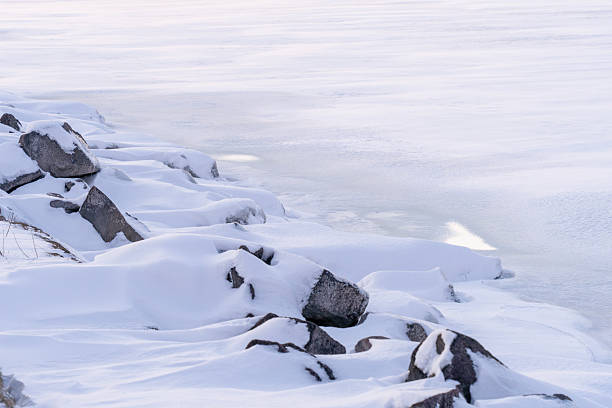 A serene winter landscape showing snow covered rocks forming a natural line along a frozen shoreline. Soft light and pale blue tones create a calm, minimalist atmosphere that emphasizes cold weather, stillness, and natural textures.