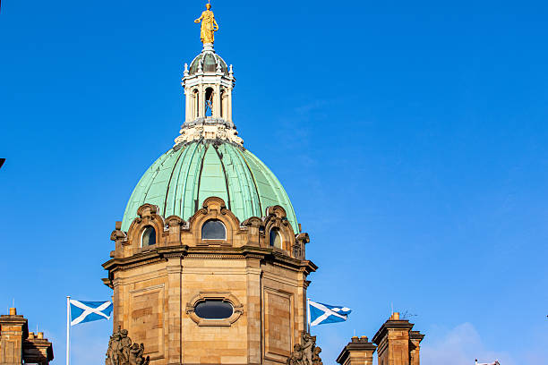 A golden statue crowns a green dome atop a sandstone building, with two Scottish flags flying against a clear blue sky  in Edinburgh, Scotland.