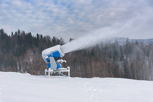 A blue mechanical snow gun blasts a thick cloud of white powder onto a winter landscape framed by a dense pine forest and a cloudy sky.