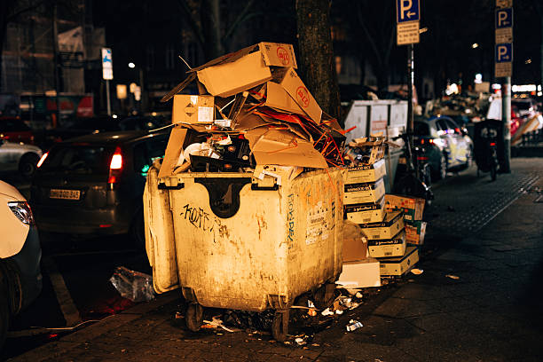 Berlin, Germany – December 21, 2025: An overflowing garbage dumpster filled with cardboard boxes and retail packaging stands along a Berlin street in the Neukölln district at night.
