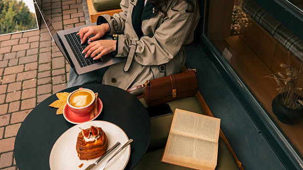A woman in a trench coat types on her laptop at a cafe table, enjoying a latte and a pastry, with a book and purse nearby