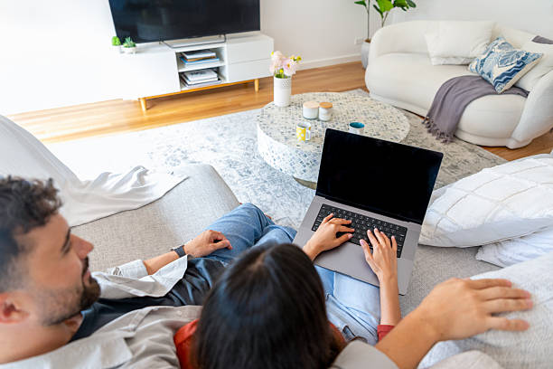 istock Couple using a laptop computer with a blank screen at home on the sofa. The house living room can be seen in the background 2256143603
