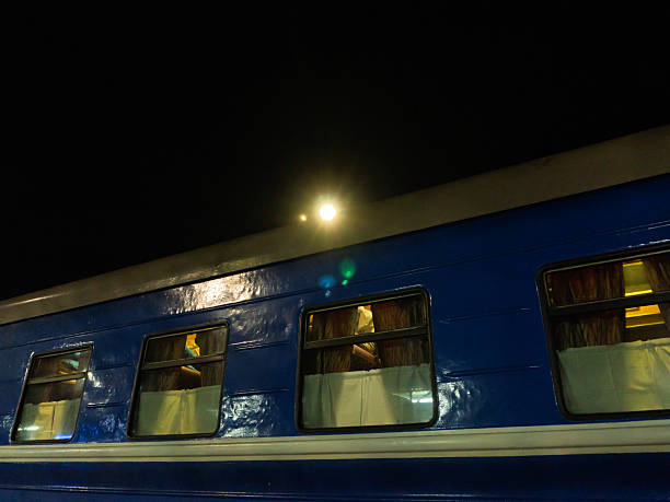 A low-angle side view of a blue passenger train carriage stationed at night. The glossy metal exterior reflects a bright light source positioned above the roofline, creating a starburst effect against the pitch-black sky. Rectangular windows reveal drawn curtains and warm interior lighting, suggesting a sleeping car or night transit. The dark background provides substantial copy space for text.