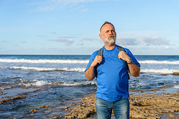 Person stands on rocky shore with backpack, facing the ocean waves under a blue sky. The sun shines down as waves crash against the rocks.