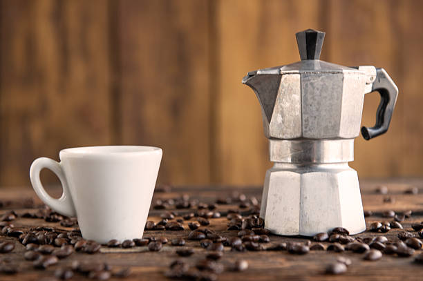 White coffee cup and moka pot placed on coffee beans against wooden background.