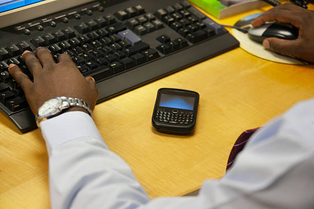 One adult African male person using a computer in an office, with a vintage-style smartphone with a physical QWERTY keyboard placed next to the keyboard