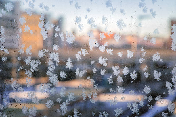 Close up view of delicate frost crystals forming natural snowflake patterns on a window glass with an out of focus urban cityscape in the background. Soft winter light and shallow depth of field create an abstract atmospheric scene symbolizing cold weather seasonal change and winter mornings in a city environment