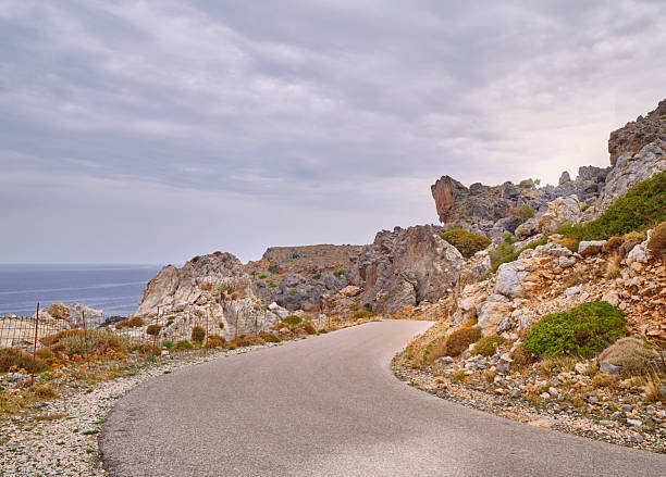 A winding coastal road on Crete, Greece curves through rocky hills with sparse vegetation, revealing the Aegean Sea on the horizon. The rugged terrain and calm atmosphere create a picturesque, travel-ready scene for landscape, adventure, and nature themes.