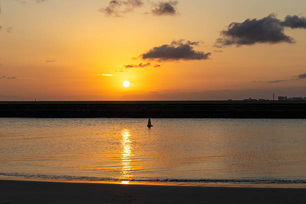 The sun sets over the third runway of Sydney Kingsford Smith Airport. In the foreground is the runway’s seawall, Botany Bay and a buoy marking the airport exclusion zone. On the right horizon are multi-storey buildings and construction cranes at Brighton Le-Sands. This image was taken from Mill Stream Lookout, Botany Bay on a hot and sunny evening at sunset on 13 January 2026.