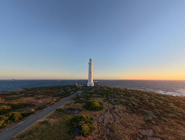 A wide panoramic view of the tallest on mainland Australia Cape Leeuwin Lighthouse perched on a rugged coastline as the sun sets, casting warm light over the  Southern and the Indian Oceans and rocky shore along the horizon.