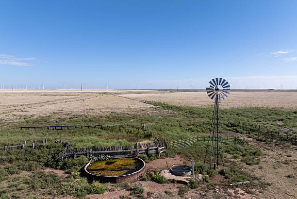 A high-angle landscape shot capturing the contrast between a vintage steel windmill water pump and a field of modern renewable energy wind turbines. Taken in the vast plains of the Texas Panhandle along historic Route 66, the image features a rustic cattle water tank, wooden fences, and a clear blue sky, symbolizing the evolution of energy and rural life in the American Midwest.