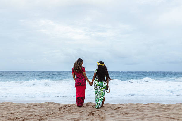 Two Marshallese young women wearing traditional-style Pacific Island dresses hold hands and look out at the ocean on a sandy beach on Oahu, Hawaii.