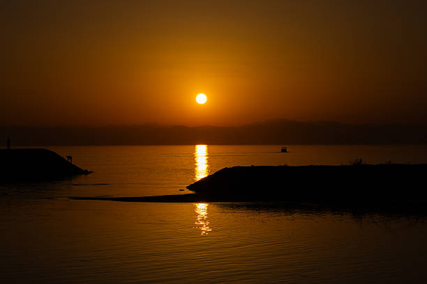 A peaceful evening scene showing a vibrant golden sunset over the Mediterranean sea with a stone pier leading into the water, creating a symmetrical and calm atmosphere.