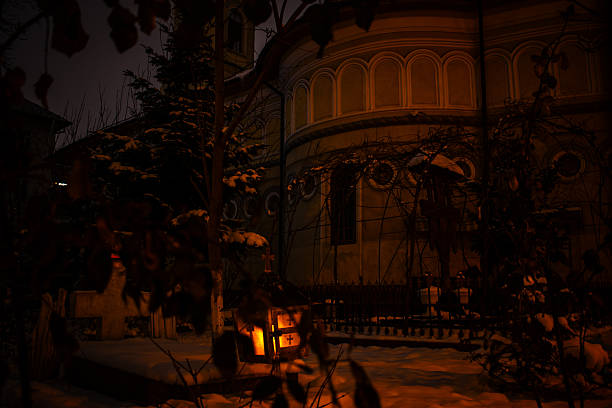 Quiet winter night scene in a historic church courtyard in Bucharest, Romania. Fresh snow covers the ground while a warm lantern glows softly near the entrance, creating a calm and spiritual atmosphere. Cinematic urban winter image with strong mood, tradition, and European character, suitable for editorial, travel, and storytelling use