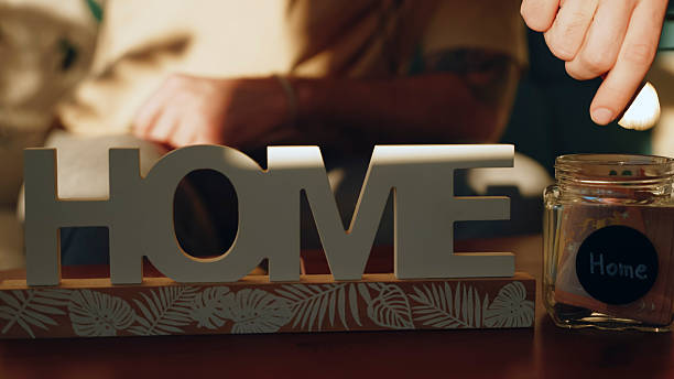 Individual placing coins into a jar labeled 'Home' for mortgage savings. Wooden 'Home' sign in the foreground. Warm lighting creates a cozy atmosphere.