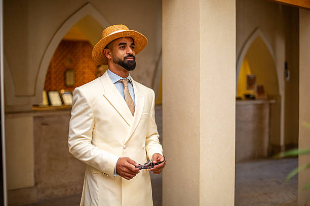 Bearded man holding sunglasses, wearing a boater hat and cream suit in an arched hallway