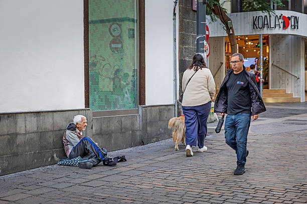 Santa Cruz, Tenerife, Spain, Europe - December 16th 2025: An elderly man is seated on the ground asking for alms in Santa Cruz, the main city of Tenerife in the Canary Islands. The picture shows a moment of social vulnerability in an urban public space, reflecting everyday realities in the city