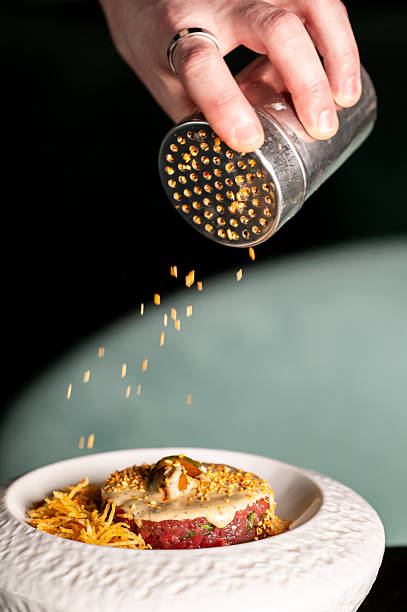 A hand uses a stainless steel shaker to sprinkle toppings over a gourmet beef tartare dish with crispy potatoes in a white bowl, capturing a culinary action shot