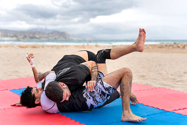 Two young adult men training brazilian jiujitsu on shore with ocean and mountains in background on an overcast day