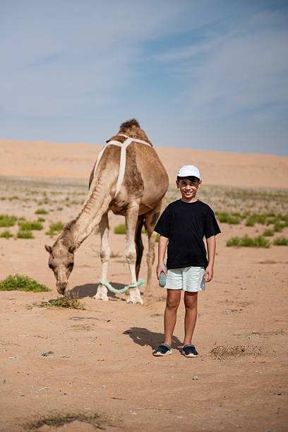 Full length side view of professional photographer and young Saudi guide in traditional attire riding camels through sandstone desert environment.