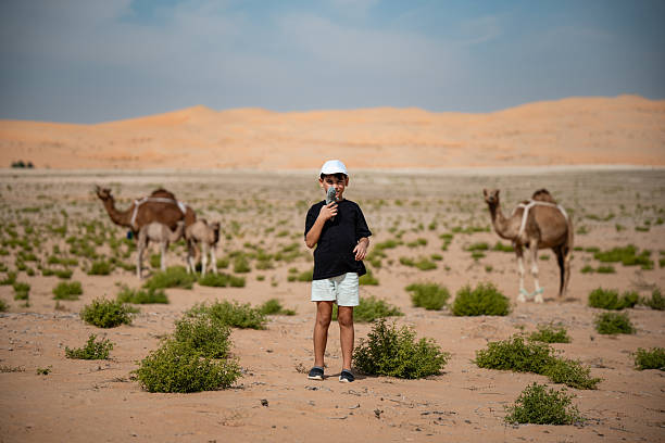 Young boy smiles holding a toy in a sparse desert landscape, with dromedary camels and calves grazing with sand dunes in background