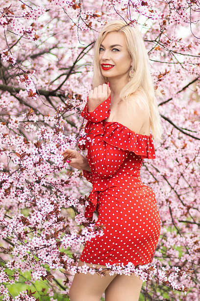 Spring beauty portrait of blonde woman in red polka dot dress surrounded by cherry blossoms. Romantic lifestyle and nature concept.