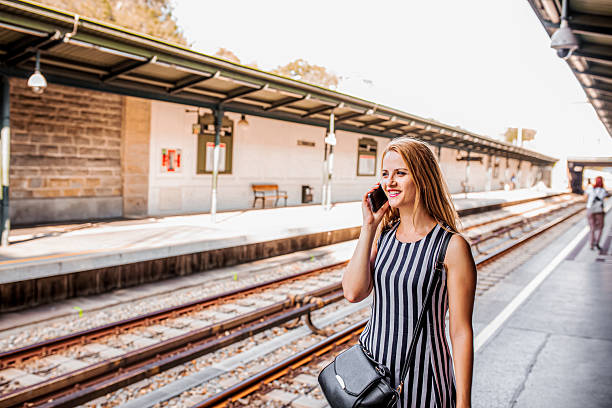 Young woman smiling, talking on smartphone while waiting for train at sunny platform