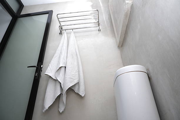 Simple modern bathroom corner with hanging towel on wall rack, white bin, and glass door, showing clean minimal interior design.