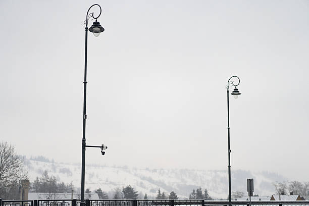 Two elegant street lights overlook a frozen valley under a grey sky while snow blankets the distant mountains and residential buildings in this peaceful scene.