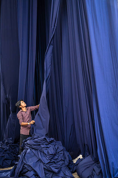A young man in a checked shirt stands amid towering panels of deep indigo fabric, pulling and arranging one of the hanging sheets. Large piles of folded cloth surround him on the ground while long vertical folds create dramatic lines and shifting shades of blue. The scene evokes a hands-on textile workshop or dyeing studio, focused on the process of handling and drying heavy bolts of fabric.