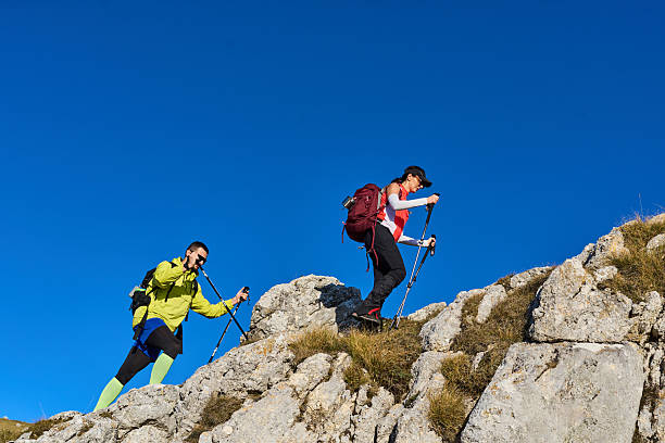 Two individuals trek up a rocky path in the mountains, using hiking poles. The sun shines brightly against a clear blue sky, creating an uplifting outdoor scene.