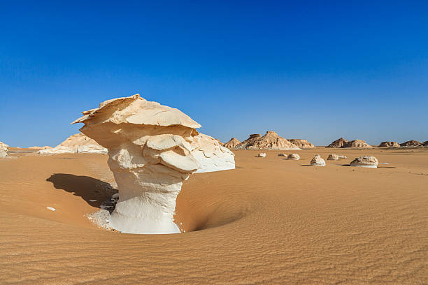 A large, white, wind‑eroded rock formation rises like a mushroom from rippled orange sand under a clear deep‑blue sky. The foreground pillar casts a sharp shadow into a shallow sand hollow, while scattered rounded white boulders and distant flattened mesas punctuate the flat desert plain.