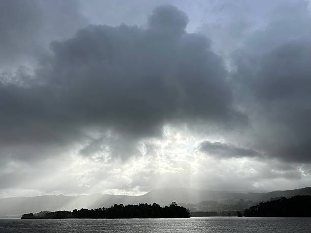 Sunlight breaks through dense storm clouds over the River Minho near Eiras, O Rosal, Galicia. The scene captures radiant beams piercing the gloomy sky, illuminating parts of the water and landscape. The river gently spans the foreground with tree-lined banks, creating a serene contrast to the dramatic sky. The distant hills are faintly visible under the cloud cover, enhancing the sense of depth and natural beauty in this tranquil setting.