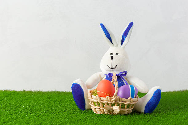 A bunny doll with a basket holds colorful eggs on grass near a plain wall.