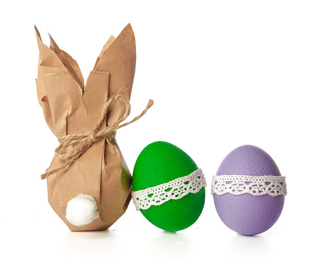 A paper bunny stands beside two painted eggs in green and purple for Easter decoration.