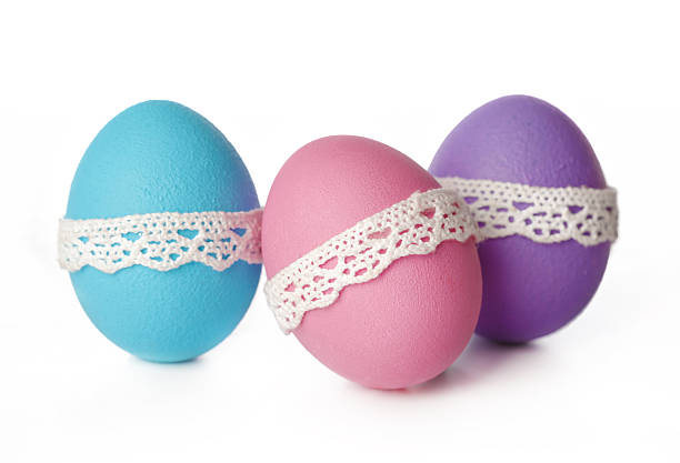 Three decorative eggs in blue, pink, and purple colors with lace bands sit on a table.