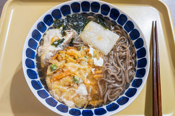 A bowl of traditional Japanese hot soba noodles topped with crispy vegetable tempura, a piece of toasted mochi, seaweed, and an egg, served in a blue patterned ceramic bowl.