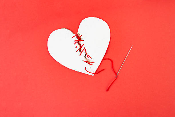 Broken cardboard heart sewed together with a needle and red thread on red background, marriage  concept