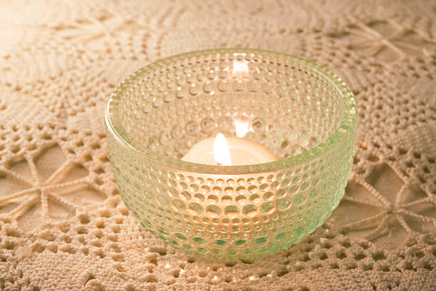 Lit candle in a textured glass holder casting a warm glow on a delicate lace tablecloth, illuminating the intricate patterns and creating a serene, peaceful ambiance