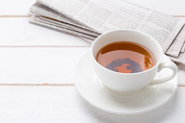 A cup of hot tea is placed on a white plate beside a stack of newspapers on a wooden table.
