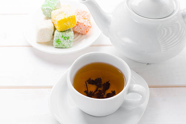 A teacup holds brewed tea next to a teapot and colored sweets on a plate.